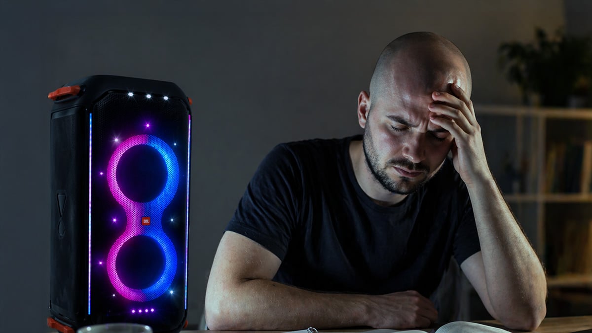 Student studying at a desk while a brightly lit speaker plays nearby, showing distraction and mental strain during focused work